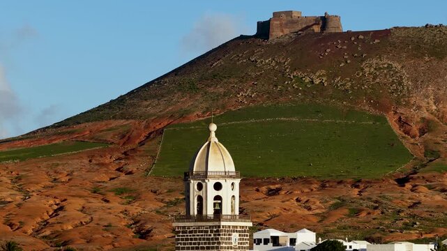 Aerial view of the iconic church steeple juxtaposed against the distant castle on the hillside, creating a striking contrast, Teguise, Canary Islands, Spain.