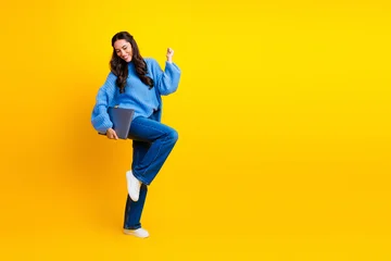 Fotobehang Onderzeeër A cheerful young mixed race woman in a blue sweater jeans and white sneakers jumps laptop yellow background conveying energy style and playfulness  © deagreez