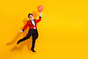 Circus magician in red suit with balloon performing joyful dance against bright yellow background at show © deagreez