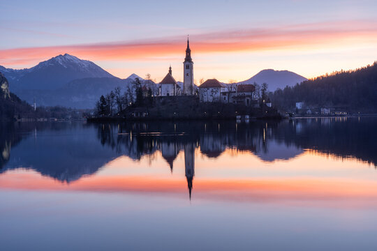 View of a church atop an island reflecting in the still waters under a vibrant sunset sky, Bled, Radovljica, Slovenia.