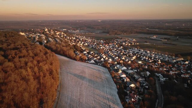 Forward-moving drone shot gliding along a forest edge over a frost-covered field, revealing Elchingen&rsquo;s village rooftops and surrounding countryside in warm golden-hour light.
