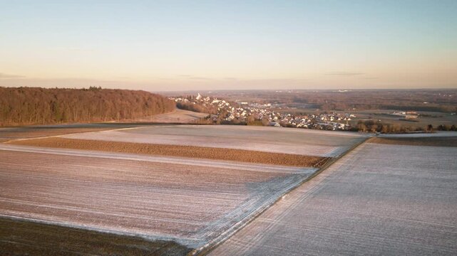 Forward-moving drone flight over frosty agricultural fields toward Elchingen, with forest edge on the left and a clear winter sky over the distant village and valley.