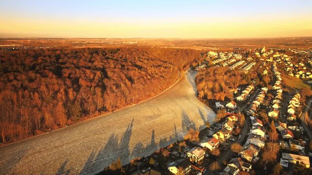 Forward-moving drone at golden hour near Elchingen, flying over a frosty field beside a dense forest and approaching residential rooftops, with warm light and long shadows.