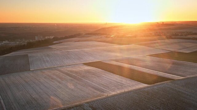 Forward-moving drone flight over frost-covered patchwork fields toward a bright sunrise, with cinematic lens flare, soft haze, and rolling farmland stretching to the horizon.