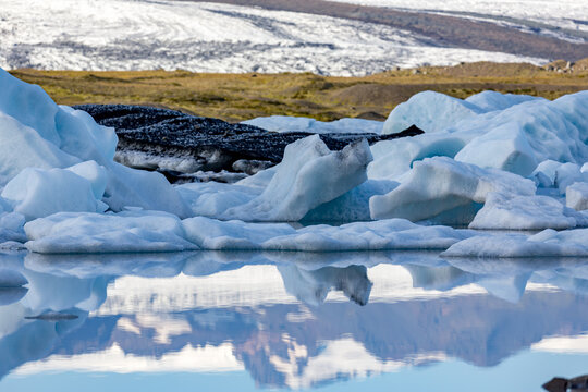 Blue and white icebergs drift in perfect reflection across J&ouml;kuls&aacute;rl&oacute;n glacier lake as soft afternoon light settles over Southern Iceland&rsquo;s rugged mountains and the distant glacier resting quietly bey