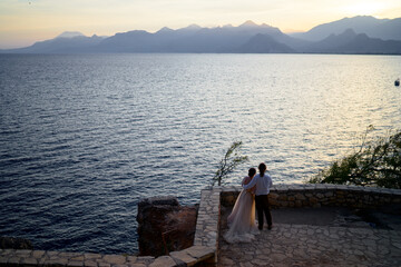 Couple stands close on a stone terrace, gazing over the calm sea as the sun sets behind distant mountains. Romantic, serene, and perfect for wedding, engagement, or travel themes. © Loginova