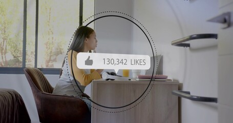 Studying female mustard-top typing on laptop at wooden desk by large window showing 130,342 LIKES