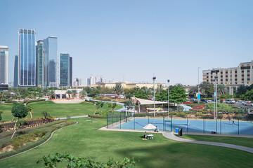 A modern urban park with green lawns, a tennis court, and skyscrapers in the background under a clear blue sky. © Loginova