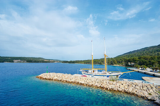 A sailboat docked at a rocky pier on a clear blue sea, surrounded by lush green hills under a partly cloudy sky.