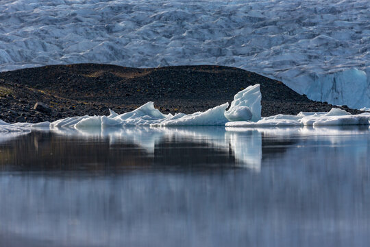 Icebergs drift in quiet harmony across J&ouml;kuls&aacute;rl&oacute;n glacier lake as soft evening light settles over Southern Iceland&rsquo;s rugged dark ridges and the distant glacier shimmering behind them