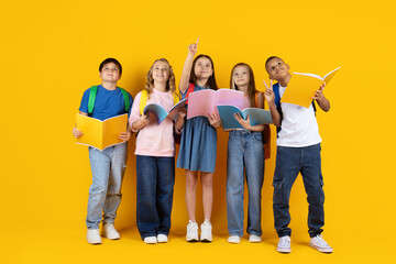 Obraz premium Preteen children looking up with books in hands on yellow studio background. Curious expressions symbolize learning, imagination, school education, and future goals