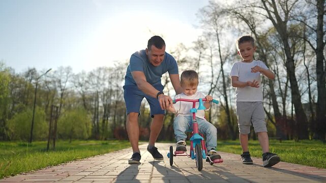 Father teaches child to ride small bike. Boy pedals along path in park. Supportive parent helps child ride. Child learns balance and movement. Boy ride with father help in outdoor park ride.
