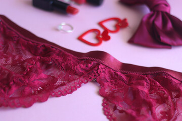 Red lace bralette, hairbow, heart shaped earrings, golden ring, red lipstick and nail polish on pink background. Coquettish accessories for Valentine's day. Selective focus.