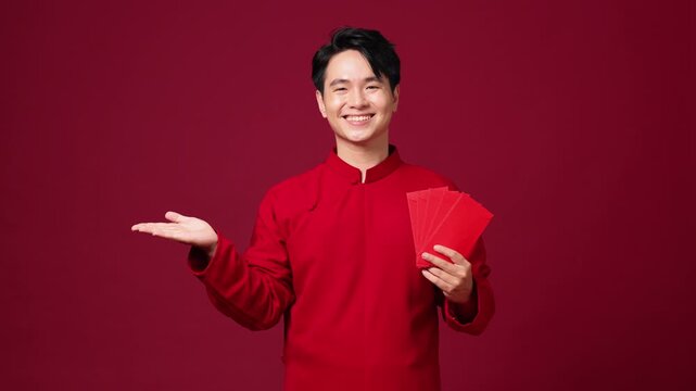 Asian man presenting with open hand gesture, holds red envelopes in another hand, red traditional outfit, promotional layout ready.