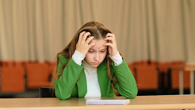 A worried schoolgirl sits with hands in her hair at a classroom table, shallow depth of field and centered framing. The image connects to test stress, learning overload, and hidden teen depression.