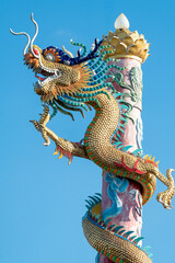 Naklejka premium Intricately detailed Chinese dragon statue with golden scales coiled around a tall red pillar. Traditional religious architecture at a temple set against a clear blue sky on a bright sunny day.