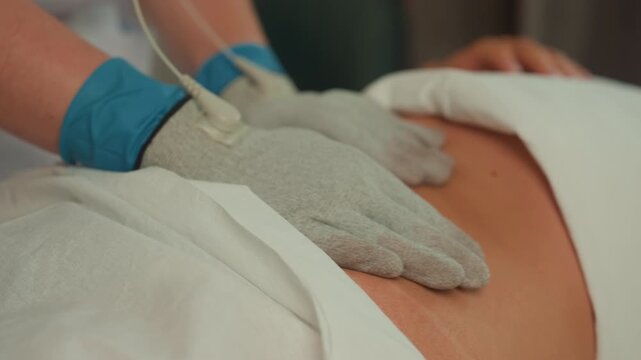 gloved hands microcurrent abdominal massage, unidentified client reclined on spa table, clinician wearing blue gloves applies conductive gloves connected to thin wires, gentle pulsed stimulation