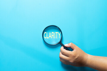 Hand holding magnifying glass focusing on the word Clarity on blue background. Business transparency, clear vision, focused mind, understanding, and simple strategy concept.