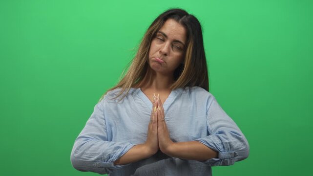 Woman with hands pressed together in prayer gesture in studio against green backdrop, pouting and rolling eyes; pleading regret.