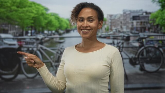 Woman smiles and points finger to canal on street in amsterdam by bicycles and bridge; confidence travel.