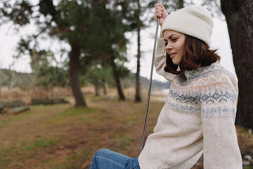 Obraz premium Woman sitting on a swing in a park, wearing a knitted sweater and beanie, relaxing outdoors. Serene casual scene with trees and soft light, perfect for lifestyle, nature, and seasonal moments.