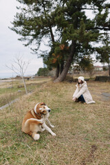 Obraz premium Woman kneeling by a calm riverside with a large dog resting on grass, trees in the background under overcast sky, a peaceful outdoor moment.
