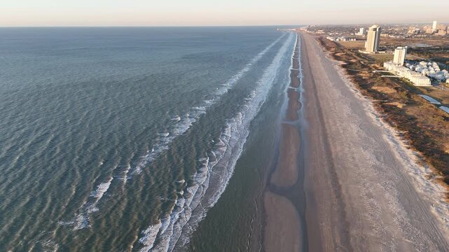 Expansive drone aerial capturing the Galveston coastline with surf on one side and wetlands, lagoons, and city buildings on the other. 