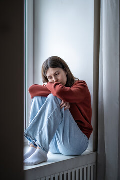Depressed lonely young woman sitting on windowsill sadly looking feeling anxiety. Alone girl suffering from emotional pain having life adolescent troubles. Mental disorder anhedonia, stress discomfort