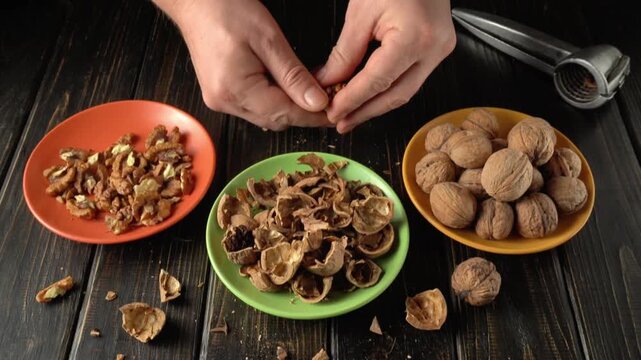 Hands of a person shelling walnuts, separating shells from nuts in colorful bowls on a wooden table with a nutcracker in the background