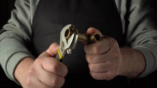 Hands of a male plumber using a wrench to tighten a brass fitting, showcasing the detailed process of plumbing work in a dimly lit workshop