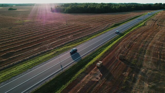 Cars drive along a country road through golden fields with harvested wheat against a warm sunset. A road through a wheat field. Aerial view from a drone. 4k footage