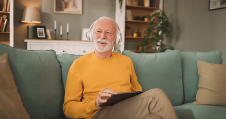 Obraz premium An elderly man with gray hair is sitting on a green couch and smiling while using a tablet in a well-lit living room. He enjoys the time alone in this space.