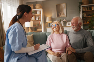 Obraz premium Home health nurse interacts with an elderly couple during a routine health check-up at their house. The couple looks engaged and comfortable in their living room while the nurse takes notes.