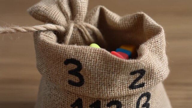 A hand reaches into a small burlap sack with numbers printed on it, pulling out colorful game pieces, suggesting a game of chance or a lottery.