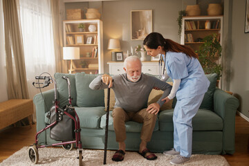 An elderly man is sitting on a couch while a home health nurse helps him stand. The nurse offers...