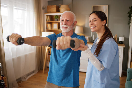 Elderly man lifts weights under the guidance of a home health nurse in a bright living room. They work together to improve strength and health.