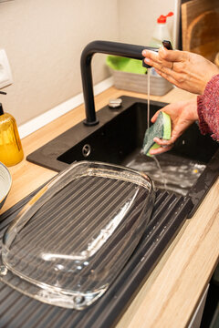 A person is washing dishes in a sink. The sink is black and has a glass dish rack underneath it. The person is holding a sponge and is washing a dish