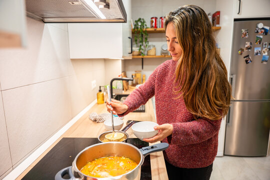 A woman is cooking in a kitchen. She is wearing a red sweater and is stirring a pot of soup