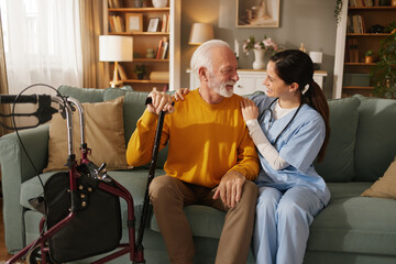 Fototapeta premium Home health nurse smiles while talking to elderly man in living room. The man uses a cane and sits on a couch. A walker is nearby as they share a moment.