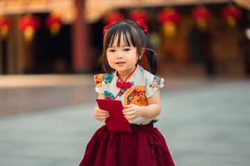 Cute Asian toddler girl in traditional Chinese dress holding a red envelope (Angpao) for Lunar New Year celebration. Chinese New Year festival. Little girl in Qipao in a temple or festive background.