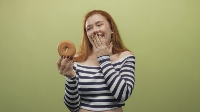 Redhead woman holding glazed donut with hand to mouth gesture in studio wearing striped off shoulder top showing midriff; guilty pleasure indulgence.