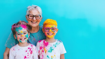 A cheerful elderly woman and two kids with colorful paint on their faces and shirts smile against a vibrant blue background.