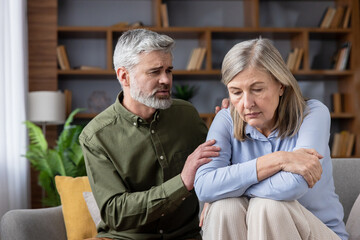 Senior man offering support and empathy to a distressed older woman, sitting together on a couch in their living room, representing companionship, care, and coping with difficult emotions