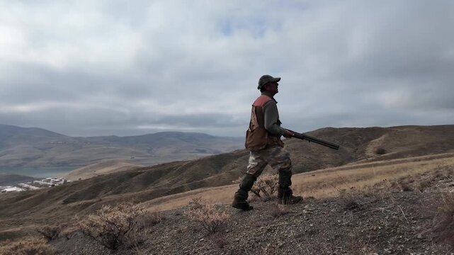 Side view of a hunter carrying a rifle while moving across a foggy mountain slope. Represents hunting, outdoor lifestyle, nature, rural culture, and documentary-style wilderness atmosphere.