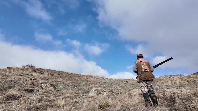 Rear view of a hunter walking through dry mountainous terrain carrying a rifle. Represents hunting, nature, outdoor lifestyle, rural environment, and exploration in a documentary-style scene.