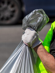 Close-up of a worker's hand in a light grey glove, securely holding the top of a large, translucent silver garbage bag.