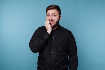 Obraz premium Man shows nervous expression while standing in front of blue background and biting his nails on a normal day at home