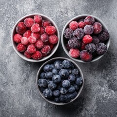 Top view of three bowls with frozen assorted berries including blueberries, raspberries and blackberries on a grey textured background