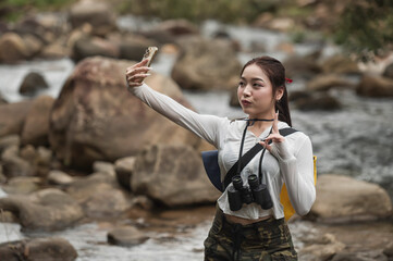 young woman hiking and selfie  in cannel. Young woman carrying a yellow backpack and camera is hiking through nature, carefully crossing a rocky stream in the forest. Outdoor adventure and eco-travel 