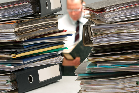Gestresster B&uuml;romitarbeiter mit Aktenordner beim telefonieren zwischen zwei Aktenstapeln.